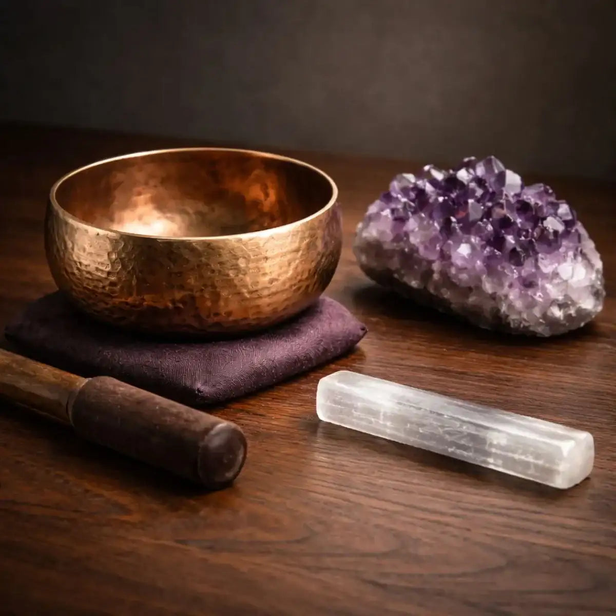 Copper singing bowl with amethyst crystal cluster and selenite wand on dark walnut surface in moody lighting